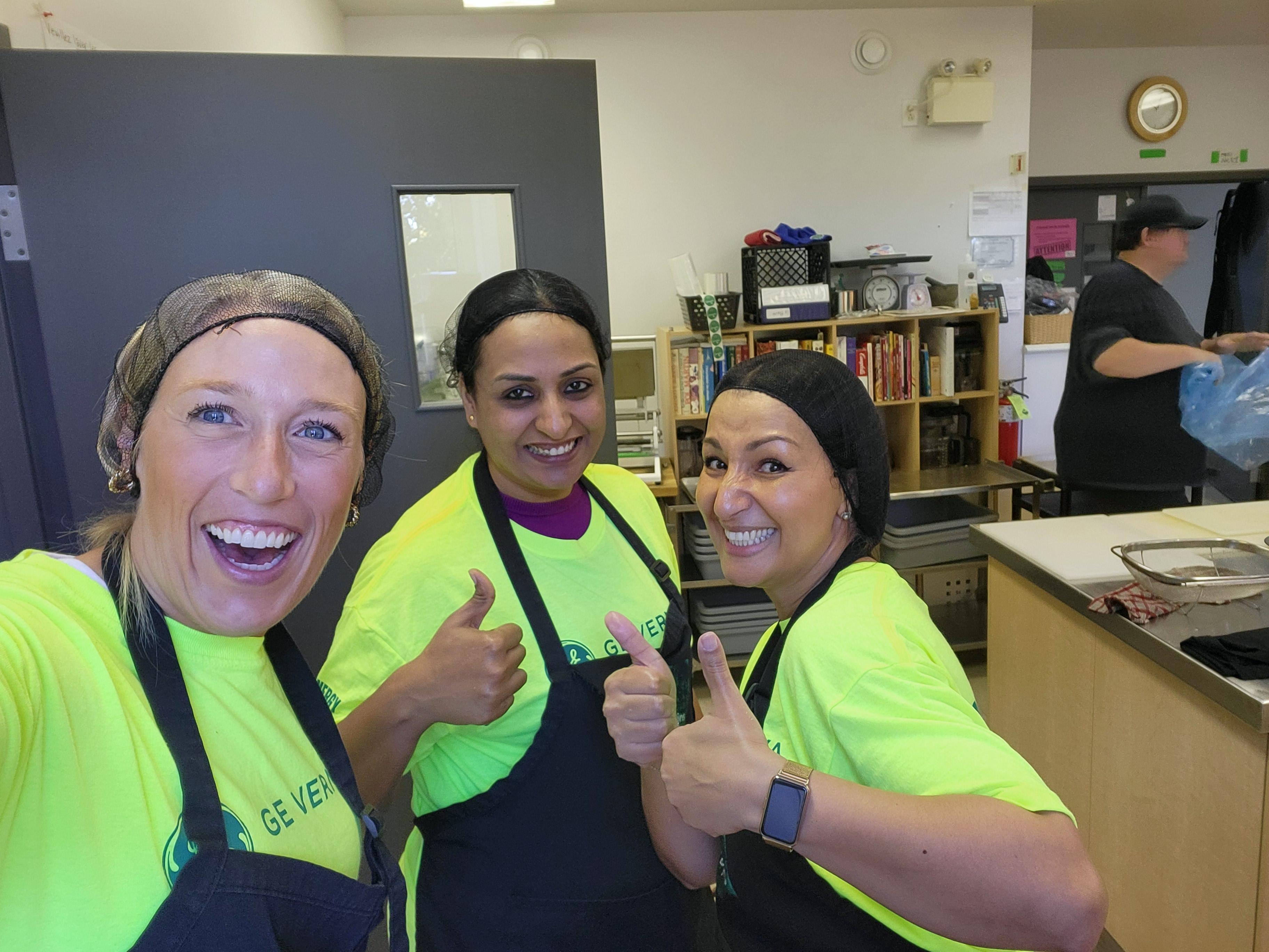 Three GE Vernova volunteers in urgency green branded t-shirts and aprons give thumbs up while serving at a community event